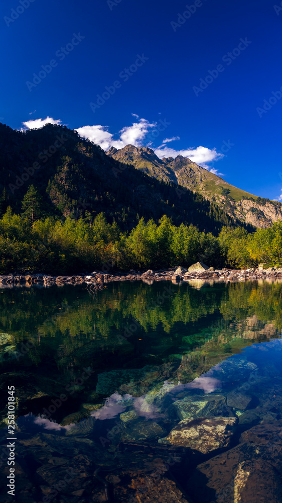 Obraz premium A huge panorama of the High-mountainous Baduk lake among the high Caucasian mountains and beautiful trees. Sunny summer day.