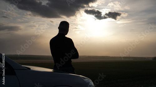 Silhouette of the young man standing near to car and looking at a sunset