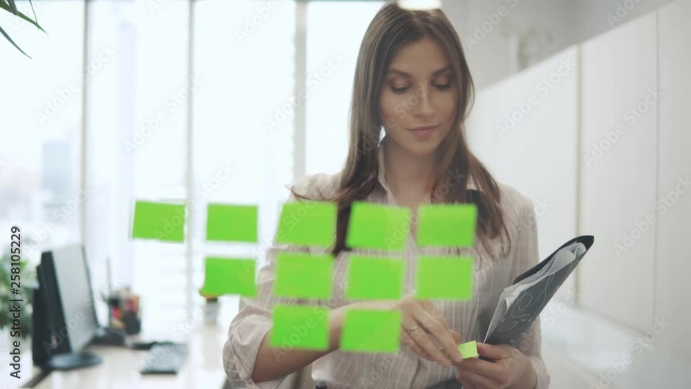 Caucasian female worker standing near glass wall with sticky notes and sticks sticky papers Caucasian female worker standing near glass wall with sticky notes and sticks sticky papers