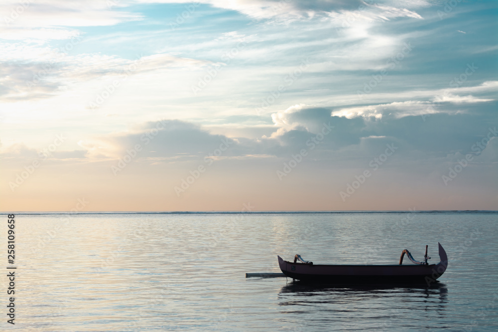 Traditional Bali Fishing Boat Without Passenger Floating On A Very Calm and Peaceful Ocean After Sunrise in Cloudy Morning Sky. Cloud Formation is Altostratus. Concept of Waiting, Peaceful and Joy Traditional Bali Fishing Boat Without Passenger Floating On A Very Calm and Peaceful Ocean After Sunrise in Cloudy Morning Sky. Cloud Formation is Altostratus. Concept of Waiting, Peaceful and Joy