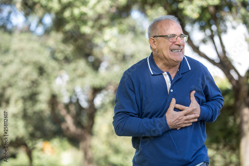 Uomo anziano con maglia blu sente un forte dolore al cuore mentre passeggia al parco