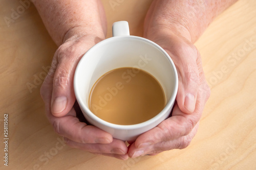 Elderly woman holds morning coffee with milk in hands, on a wooden table