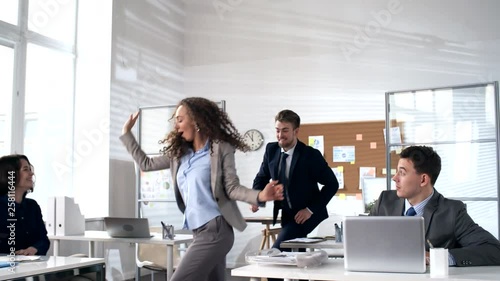 Young energetic businesswoman walking to colleagues sitting at their desks in office and inviting them to dance together: team joining her and dancing in conga line with full energy photo