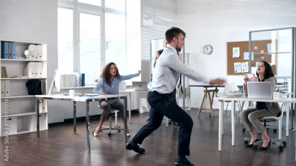 Young excited businessman dancing and performing acrobatic tricks in the center of office while cheerful colleagues smiling and photographing him with smartphones Young excited businessman dancing and performing acrobatic tricks in the center of office while cheerful colleagues smiling and photographing him with smartphones