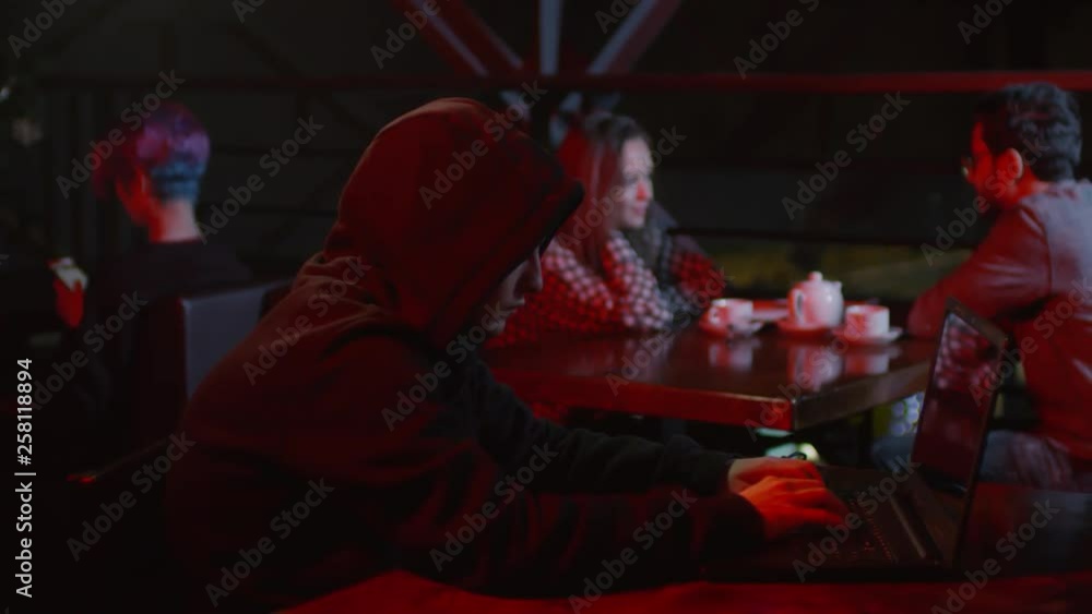 Medium shot of dangerous hooded hacker sitting at bar table at laptop and programming Medium shot of dangerous hooded hacker sitting at bar table at laptop and programming