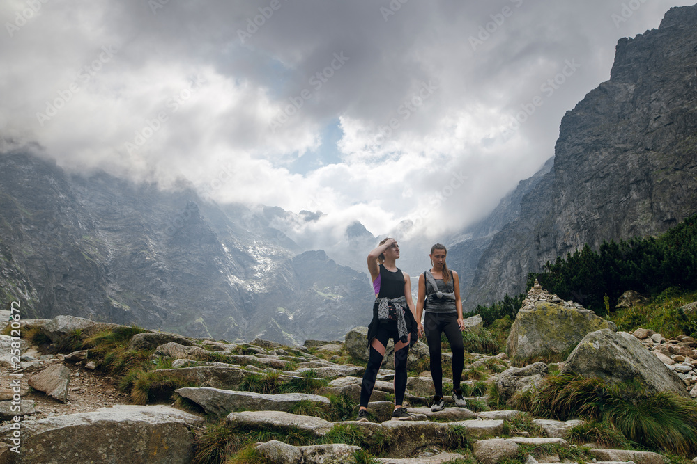 Two young women friends in sportswear hiking on the stony pathway, and cliffs in cloudy mountains. Morskie Oko lake, High Tatras, Zakopane, Poland.