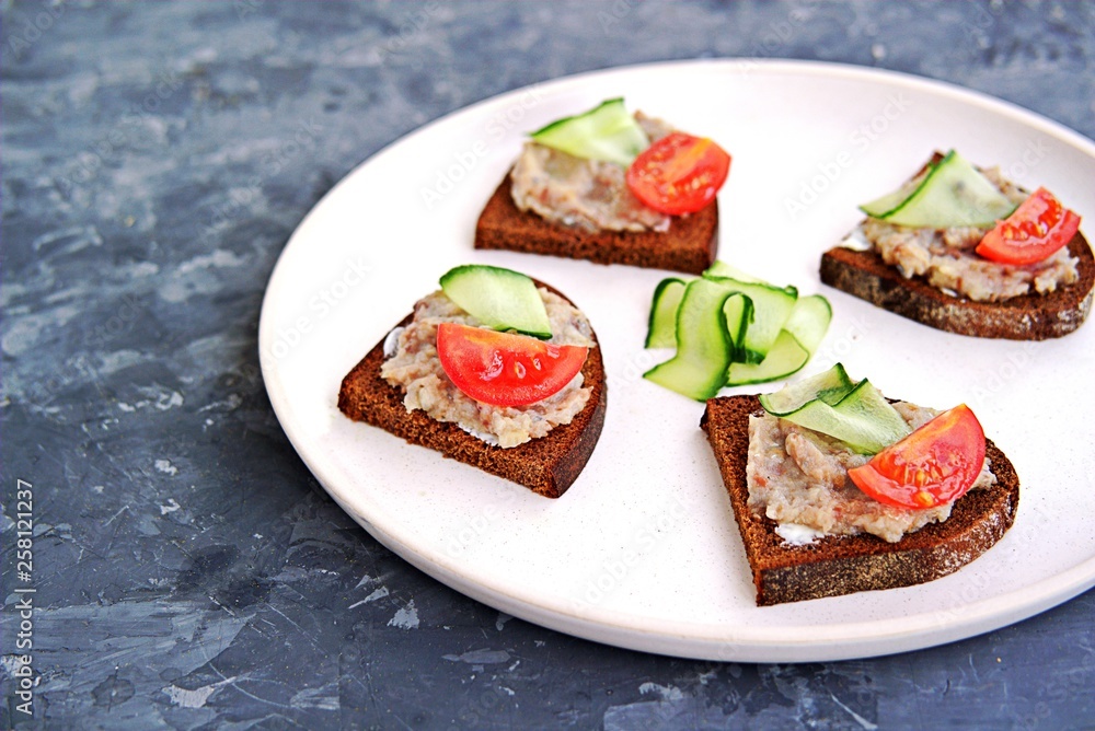 Open sandwiches on rye bread with forshmak, a snack of chopped salted herring, onions and eggs on a ceramic plate. Decorated with sliced fresh cucumbers and tomatoes.