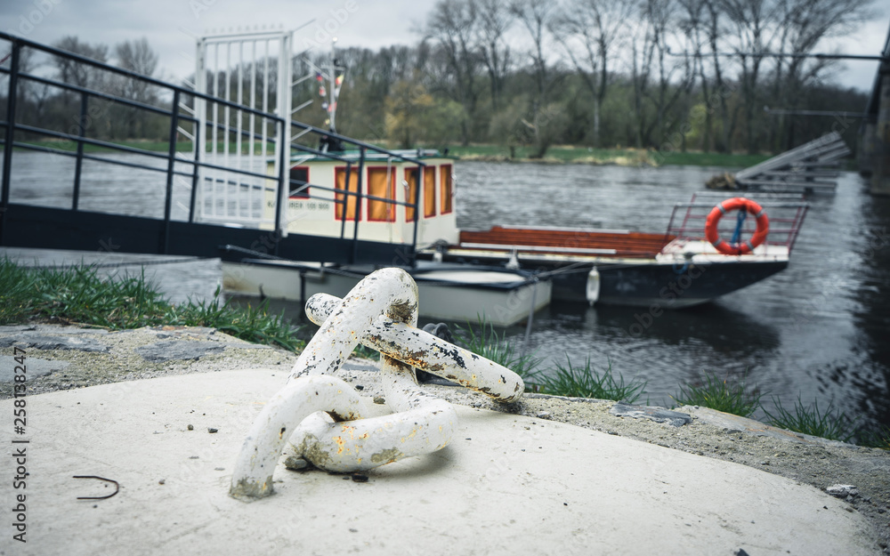 Fototapeta premium Iron old anchor with rope by the river. Ship's knot. In the background a small blue pier and an old shipping boat