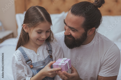 Bearded long haired happy man smiling, giving small gift to his cute little daughter. Adorable little girl receiving a present from her loving father. Celebrating, birthday concept