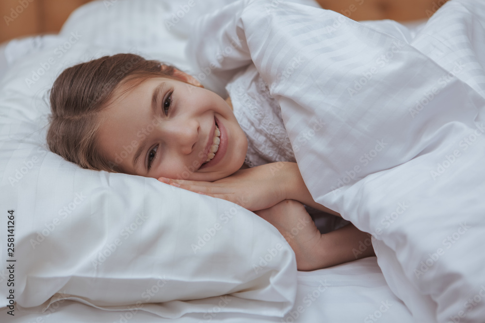 Lovely little happy girl laughing excitedly to the camera, lying in her bed in the morning. Adorable little girl waking up, smiling to the camera. Children, family, happiness concept