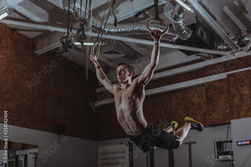 Low angle shot of a male crossfit athlete exercising on gymnastic rings. Shirtless strong sportsman working out on gymnastic rings at crossfit box gym © mad_production