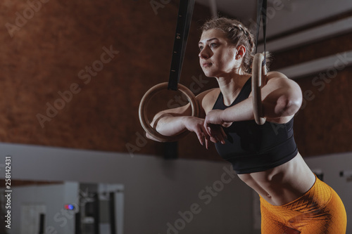 Low angle shot of a young athletic woman looking away thoughtfully, resting after working out on gymnastc rings. Female crossfit athlete relaxing after exercising, copy space