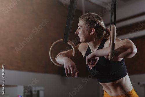 Happy healthy athletic woman laughing, looking away, resting after gymnastic rings workout. Cheerful crossfit female athlete relaxing at the gym, after exercising