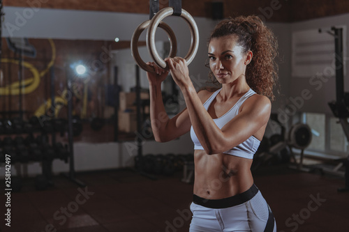 Gorgeous athletic woman with perfect abs looking to the camera fiercely, resting after gymnastic rings workout. Beautiful sportswoman relaxing at crossfit box gym, copy space