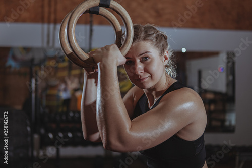 Charming female crossfit athlete smiling to the camera, holding gymnastic rings. Sportswoman smiling joyfully relaxing at crossfit gym after exercising, copy space