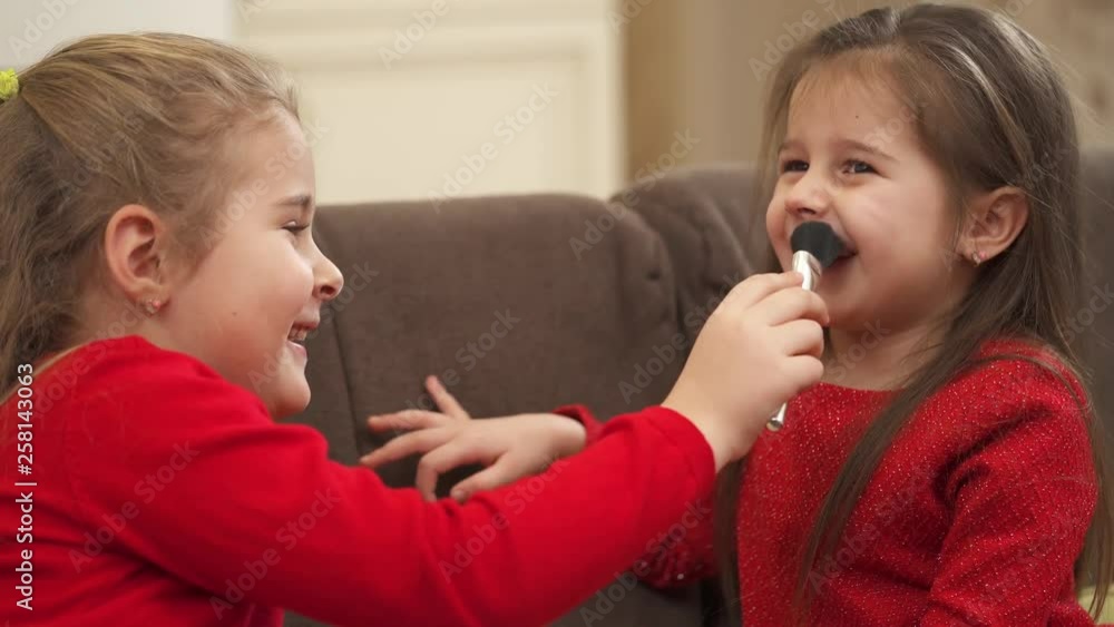 Little girl playing with a makeup brush and tickling her friend's nose with it Little girl playing with a makeup brush and tickling her friend's nose with it
