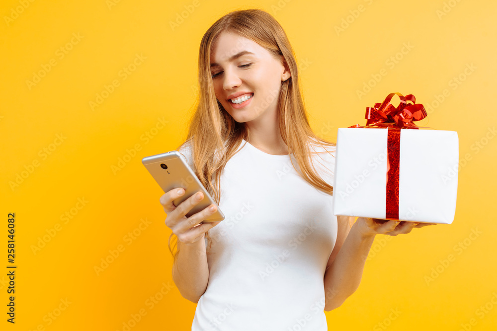 Portrait of smiling attractive woman, holding gift box, standing and using mobile phone, isolated on yellow background Portrait of smiling attractive woman, holding gift box, standing and using mobile phone, isolated on yellow background
