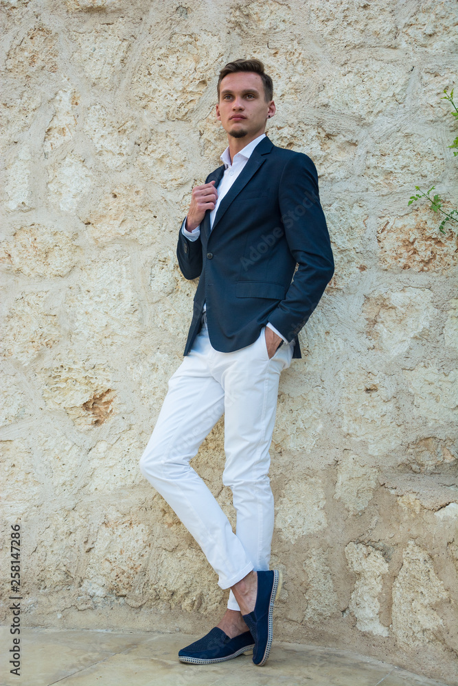 Young handsome man in a suit and white trousers at standing near a stone wall.