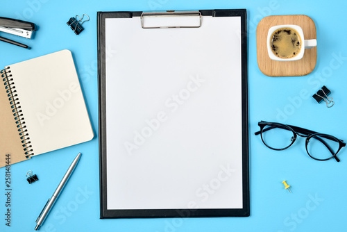 Mock up workspace with blank clip board, office supplies, pen, coffee cup, notepad, stapler and eyeglasses on blue background. Flat lay, top view, copy space