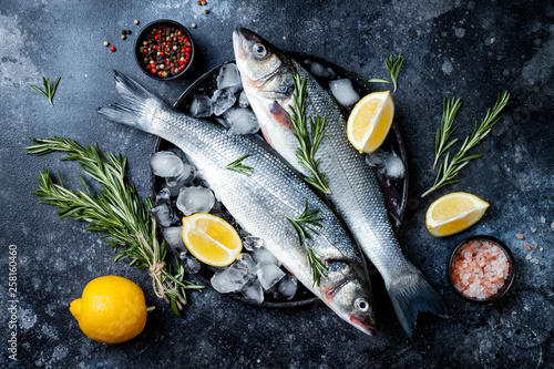 Canvas Print Fresh raw seabass fish on black stone background with spices, herbs, lemon and salt