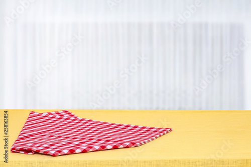Empty bright table with folded red checkered tablecloth in front of blurred curtain window natural bright background. Template for your product display montage. Space for design.