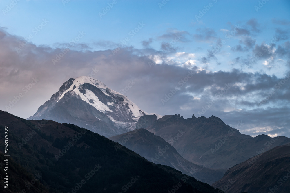 Dark rocks and snow-capped Kazbek mountain against backdrop of gloomy sky. Caucasian mountains. Landscape.