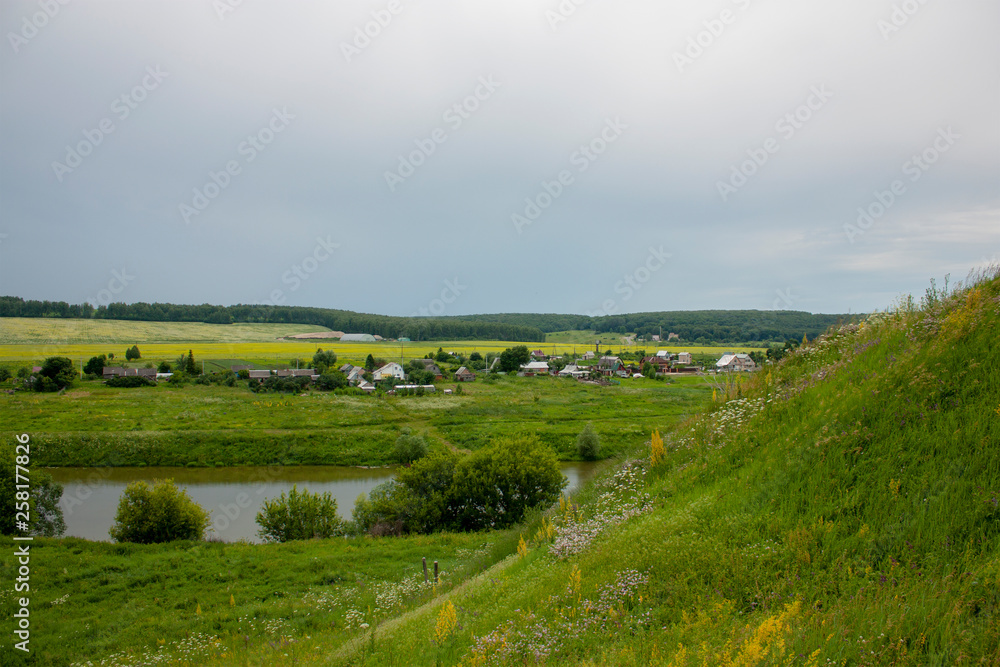 View on the settlement near the river from the hill. Green meadows and trees in the summer in Russia.