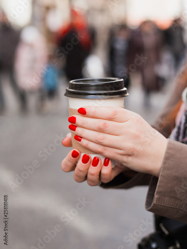Woman holding coffee cup on the blurred street. Sunny spring afternoon. Red manicure