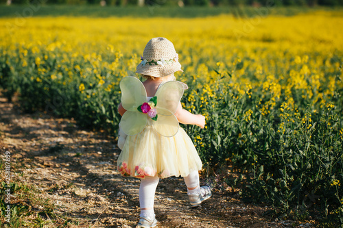 cute toddler girl walking in yellow fairy dress and straw hat in rapes field, summer, sunset. View from back.