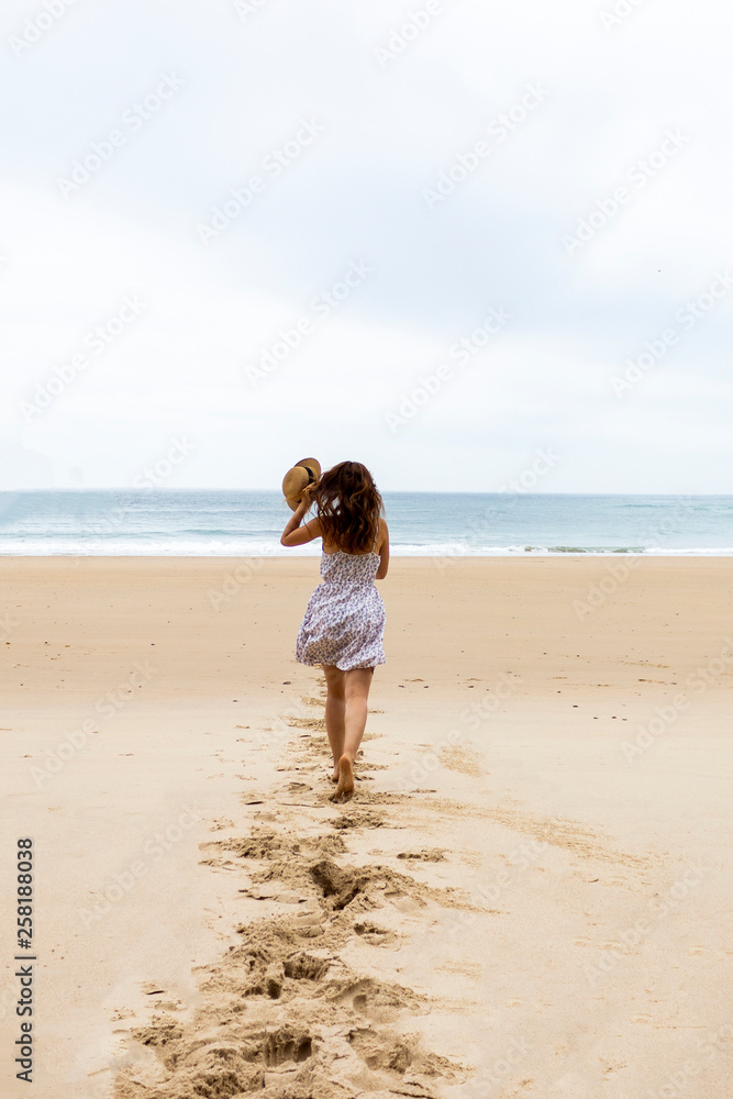 Girl in hat and white dress running towards water on the empty beach