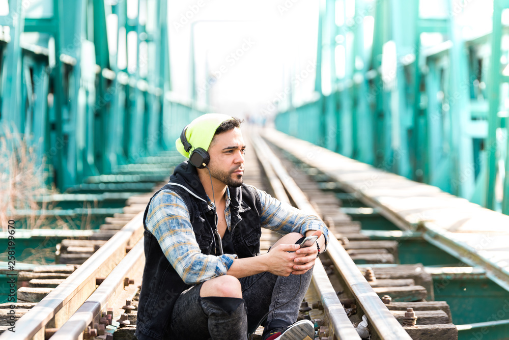 Handsome Hipster Guy Using the Cellphone and Wearing Headphones. Sitting on the Train Tracks.
