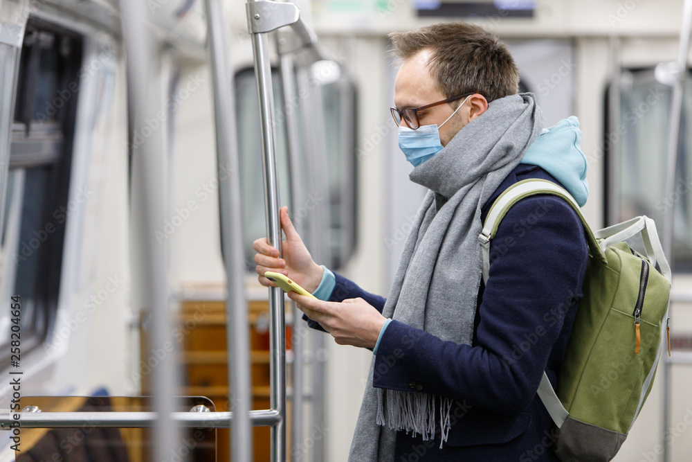 Ill man in glasses feeling sick, wearing protective mask against transmissible infectious diseases and as protection against the flu in public transport/subway,  using and looking at smartphone