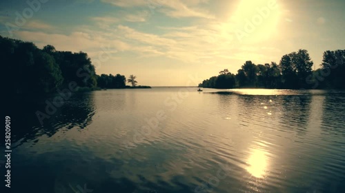 In the middle of a wide river a fisherman catches a boat. The bright sun shines in the blue sky. Along the banks are green trees and dense forest.