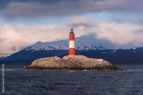 Typical view at sunset of Les Eclaireurs lighthouse, wrongly known as End of the World lighthouse. With red and white stripes on a rocky island in Beagle Channel, with clouds and mountains. Ushuaia