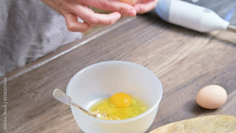 Close-up of hands. The girl at the home kitchen breaks chicken eggs into a plastic bowl against the background of a blender lying on the table Close-up of hands. The girl at the home kitchen breaks chicken eggs into a plastic bowl against the background of a blender lying on the table