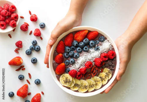 Woman hands holding an Acai Berry superfoods smoothies white bowl with chia seeds, strawberry, goji berry, coconut, blueberry, raspberry toppings, and chocolate chip on white background