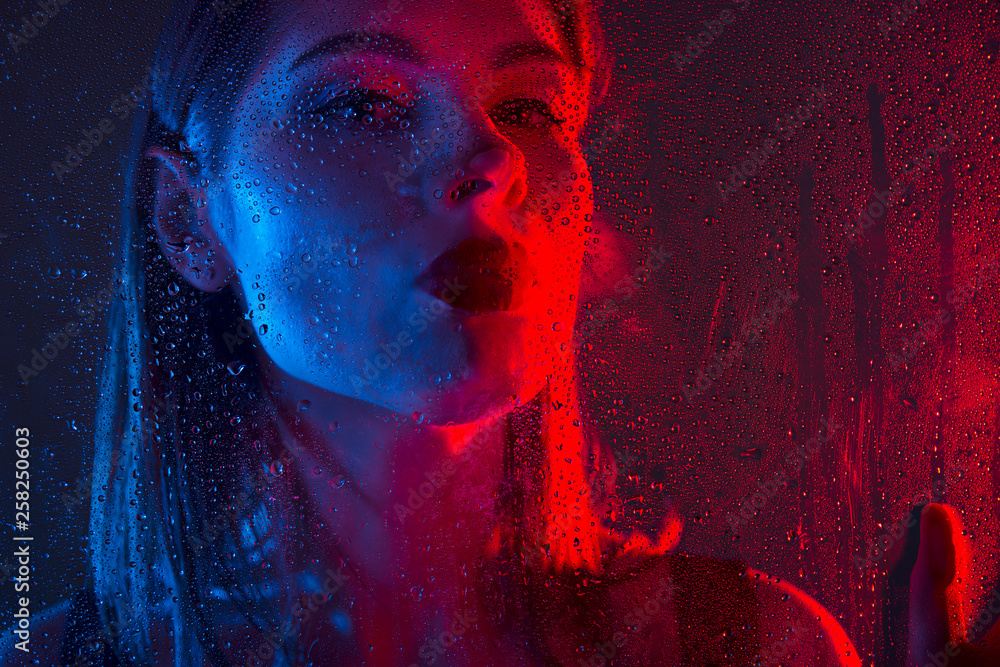 Glamorous brown-haired woman breathes on a window with water drops in neon light close-up. Glamorous brown-haired woman breathes on a window with water drops in neon light close-up.
