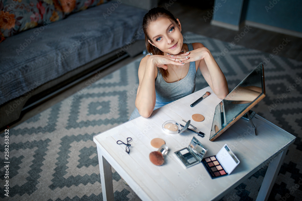 Young woman makes make-up before mirror. Cosmetics, brushes and sponges on the white table. Woman looks in the mirror. Young woman makes make-up before mirror. Cosmetics, brushes and sponges on the white table. Woman looks in the mirror.
