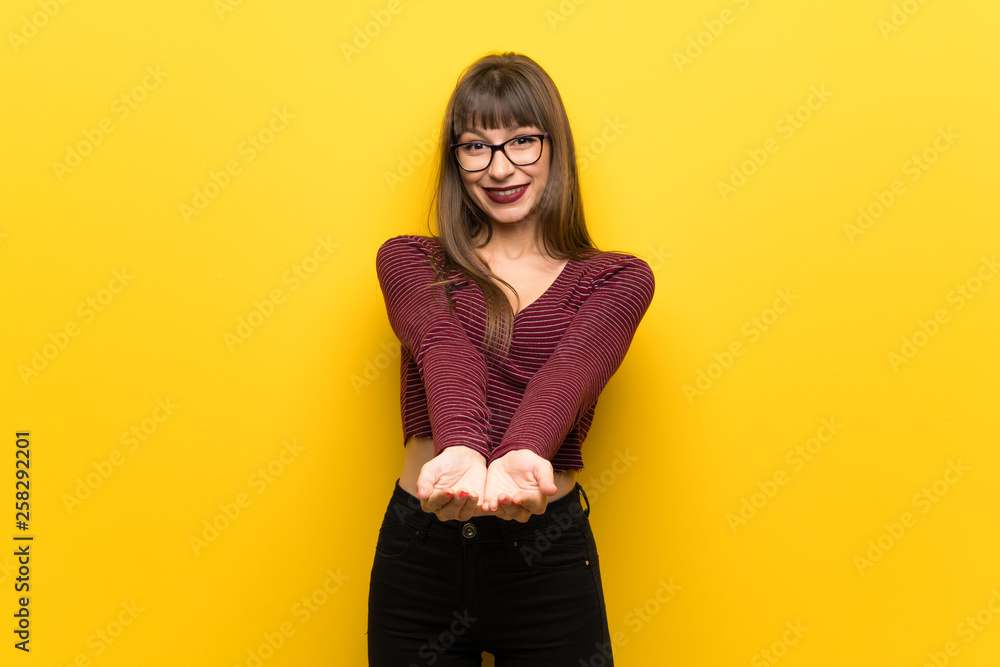 Woman with glasses over yellow wall holding copyspace imaginary on the palm to insert an ad Woman with glasses over yellow wall holding copyspace imaginary on the palm to insert an ad