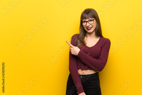 Woman with glasses over yellow wall pointing to the side to present a product