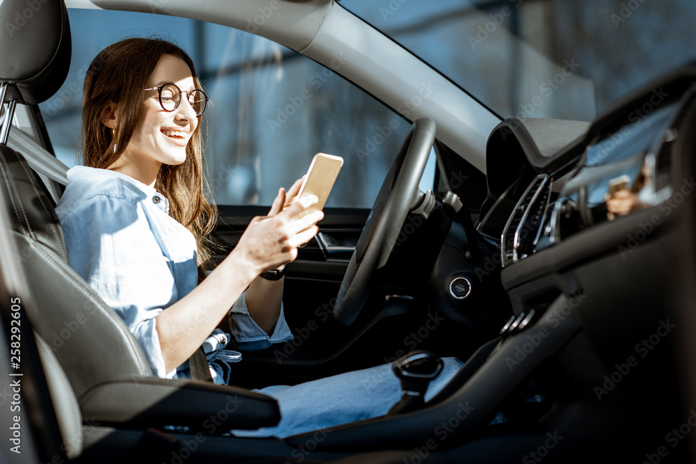 Young and cheerful woman using smart phone while sitting in the modern car in the city