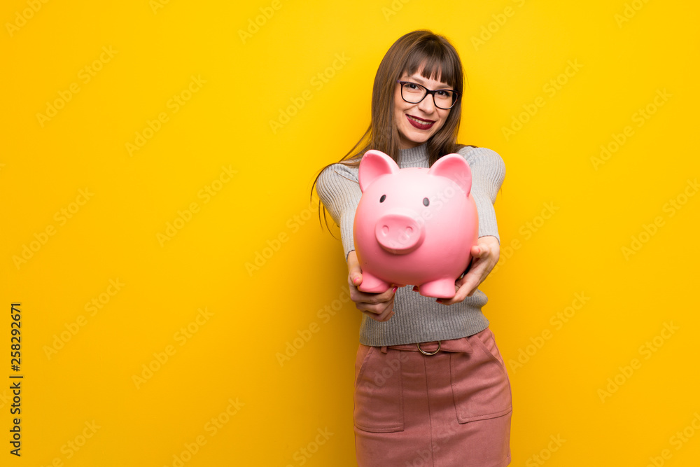 Woman with glasses over yellow wall holding a piggybank Woman with glasses over yellow wall holding a piggybank
