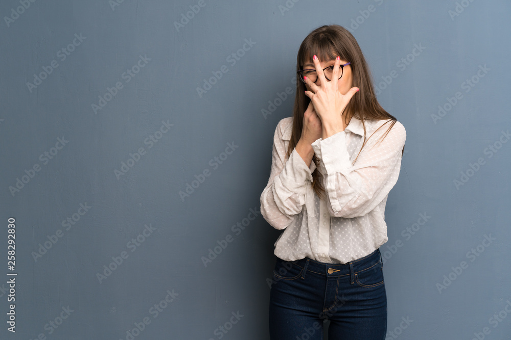 Woman with glasses over blue wall covering eyes by hands and looking through the fingers Woman with glasses over blue wall covering eyes by hands and looking through the fingers