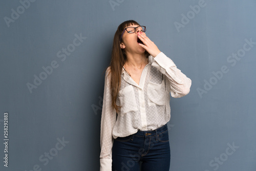 Woman with glasses over blue wall yawning and covering wide open mouth with hand © luismolinero