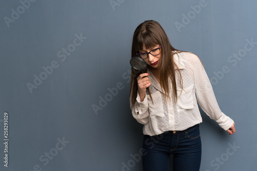 Woman with glasses over blue wall holding a magnifying glass photo