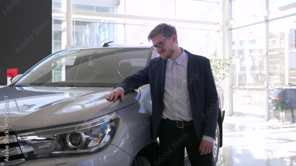 auto purchase, young buyer man with glasses gladly strokes new car and gives positive gesture in showroom auto purchase, young buyer man with glasses gladly strokes new car and gives positive gesture in showroom