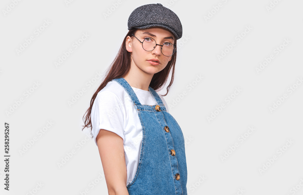 Studio image of pretty young woman wears transparent trendy eyewear, casual white t-shirt, denim dress, trendy gray cap, looks seriously directly into camera, poses on white wall. People emotions Studio image of pretty young woman wears transparent trendy eyewear, casual white t-shirt, denim dress, trendy gray cap, looks seriously directly into camera, poses on white wall. People emotions