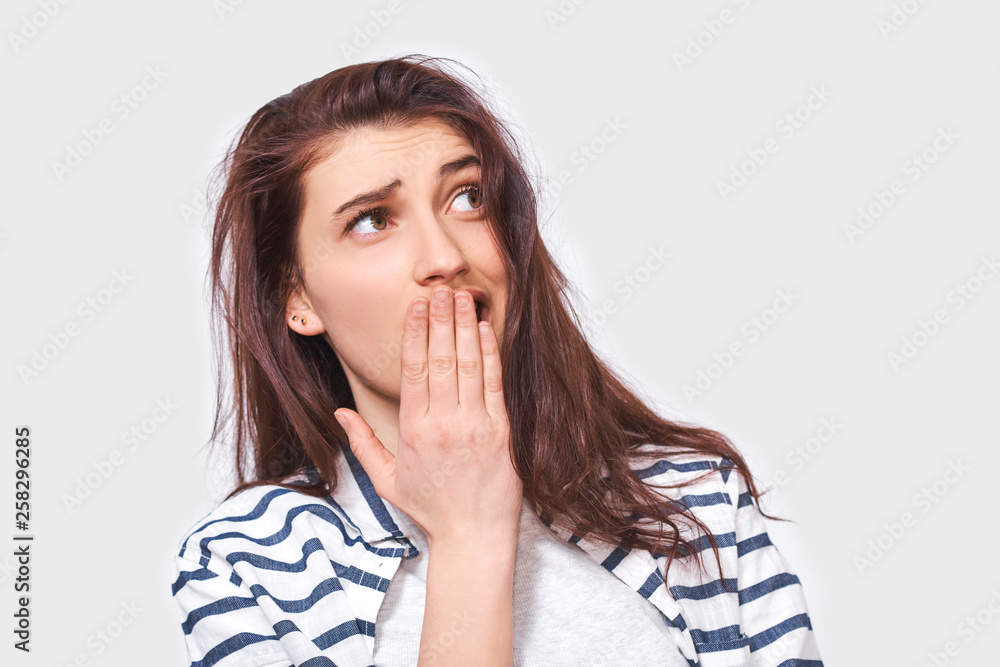 Surprised pretty young woman wears casual striped shirt, looking up to blank copy space, isolated on white studio background. Surprised girl with hand on mouth has surprised expression. Surprised pretty young woman wears casual striped shirt, looking up to blank copy space, isolated on white studio background. Surprised girl with hand on mouth has surprised expression.