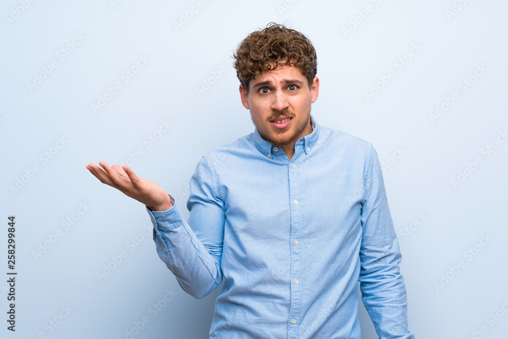 Blonde man over blue wall making doubts gesture Blonde man over blue wall making doubts gesture