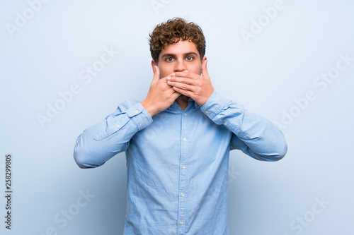 Blonde man over blue wall covering mouth with hands © luismolinero